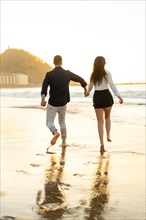 Vertical rear view of a romantic couple playing with sea water running holding hands during sunset