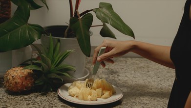 A hand poking a piece of chopped pineapple pieces on a plate with a fork for a healthy vegan summer