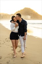 Vertical photo of a caucasian young couple kissing while walking barefoot along a beach during