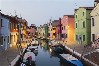 Moored boats on canal lined with colourful stucco houses, shops and footbridge at dusk, Burano
