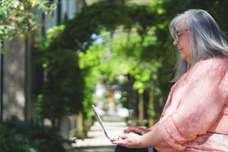 Older woman with white hair and glasses sitting on a park bench working with her laptop computer
