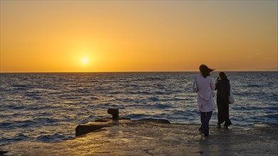 A couple stands on the shore chatting with a view of the sun setting over the sea, dusk, sunset,