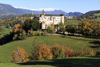 A castle surrounded by autumn trees and green meadows against a mountain background, Italy,