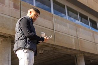 Young businessman walking down the street holding cell phone and glass of coffee, A latin man