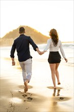 Vertical photo of the rear view of happy couple holding hands running along a beach during sunset