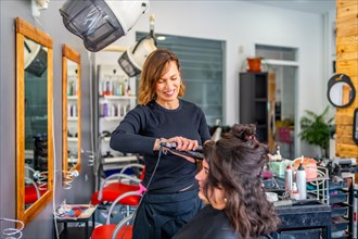 Smiling female hairdresser flat ironing hair of a woman sitting in front of a mirror in the salon