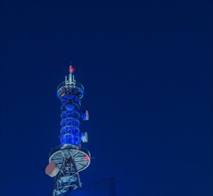 Low angle night view of large communication array tower lit up in blue lights with dark blue sky in