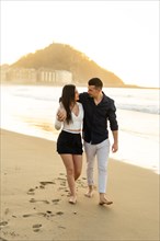 Vertical photo of a young chic couple walking barefoot along the beach during sunset