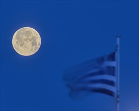 A full moon next to a waving Greek flag in the blue hour, twilight, Mandraki Oat, Rhodes,