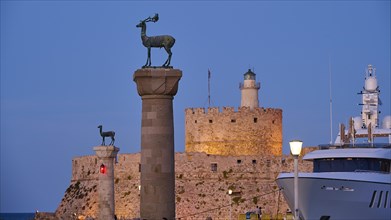 A deer sculpture on a pillar in front of a castle and lighthouse next to a luxury yacht at dusk,