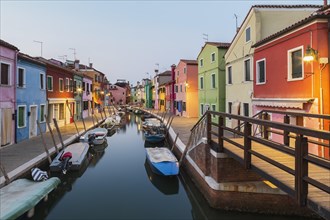 Moored boats on canal lined with colourful stucco houses, shops and footbridge at dusk, Burano