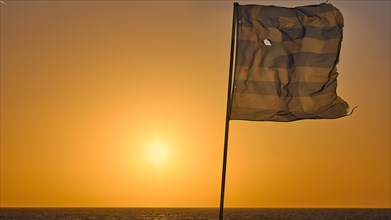 A Greek flag fluttering in the wind in front of a bright orange sunset over the sea, dusk, sunset,