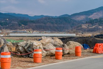 Orange traffic barrels on side of rural road in front of large boulders at construction site in