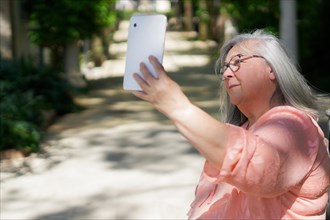 Positive old lady taking selfie in park. Senior grey haired woman in casual holding gadget with