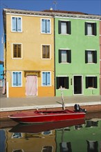 Moored red boat on canal lined with orange and green stucco houses decorated with striped curtains