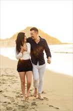 Vertical photo of a romantic smiling couple walking together barefoot along a beach during sunset