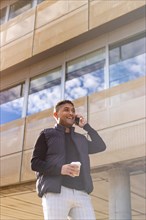 Young smart casual businessman walking down the street talking on the phone holding glass of coffee