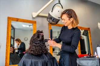 Smiling hairdresser curling the hair of a client sitting next to a mirror in the salon