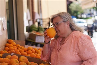 Older white-haired woman smelling an orange in a street market