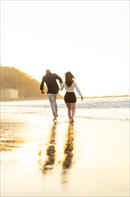 Vertical photo of a young elegant couple running holding hands along the beach during sunset