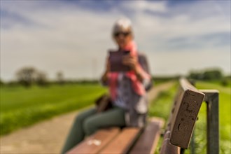 Senior citizen sitting on a bench and looking at her smartphone, side view, blurred, dike crest,