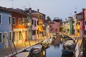 Moored boats on canal lined with colourful stucco houses and shops at dusk, Burano Island, Venetian