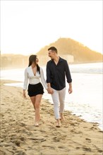 Vertical photo of a young caucasian chic couple walking barefoot holding hands along a beach during