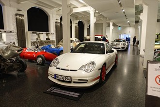 A white sports car stands in the foreground of a historical vehicle exhibition in a museum,