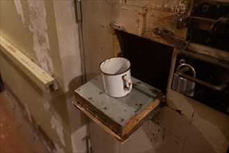 Rusted metal cup stands on the food hatch of a prison cell, former headquarters and prison of the