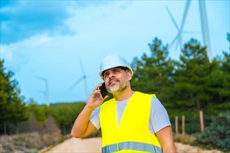 Smiling worker in reflective waistcoat and helmet using phone in a wind energy park