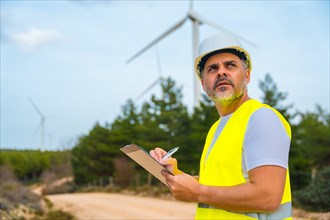 Worker in protective clothes inspecting wind turbines writing notes stranding outdoors