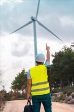 Vertical photo of the rear view of a male caucasian adult engineer pointing up to a wind turbine
