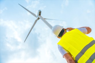 Low angle view photo of a male caucasian adult Engineer using phone next to a wind turbine in a