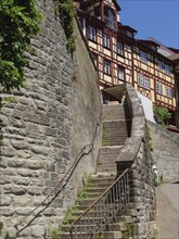 A steep staircase leads next to a high stone wall to a half-timbered house in a sunny old town,
