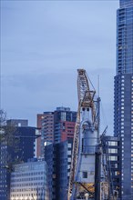 A construction crane in front of skyscrapers in a modern cityscape under a cloudy sky, small