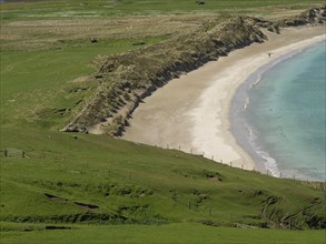 A stretch of coastline with a curving sandy beach surrounded by green meadows and rolling hills