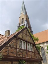 A half-timbered house in front of a high church tower, cloudy sky and a red tiled roof, large