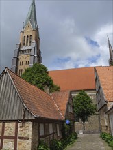 Older half-timbered houses in front of a high gothic church tower with trees and cloudy sky, large