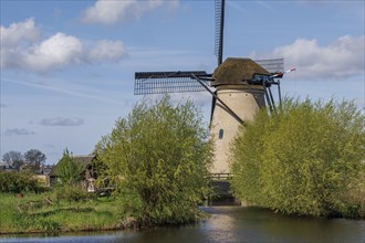 A windmill stands against a clear blue sky, surrounded by lush greenery and water, many historic