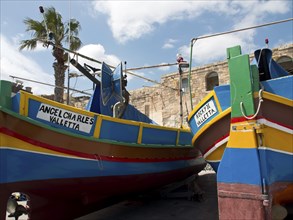 Two colourful boats on land, next to a palm tree and an old building with blue sky, many colourful
