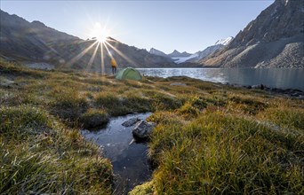 Young man with green camping tent in the mountains, mountains reflected in the lake, sunrise at