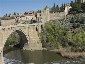 Stone bridge over a river in front of a castle wall and a city view with vegetation, blue sky,