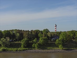 A red and white lighthouse in the middle of a wooded landscape, with the river in the foreground,