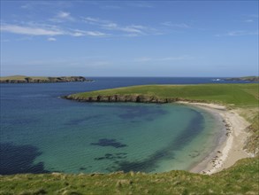Small bay with clear water and a sandy beach surrounded by green grassland and a wide blue sky,