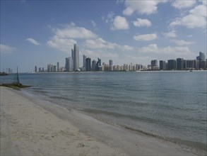 A wide sandy beach with a view of modern skyscrapers of a city by the sea under a cloudy sky,