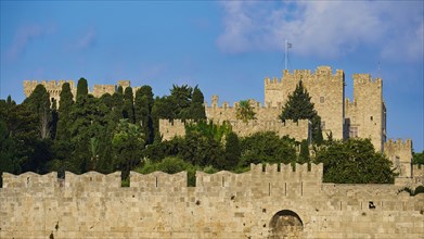 Fortress walls with battlements under a bright blue sky, surrounded by green vegetation, part of