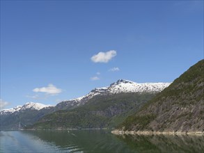 A clear lake surrounded by snow-capped mountains and a bright blue sky, calm water in a fjord with