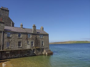 Large stone building on the edge of a sandy beach and clear blue water with a blue sky, old stone