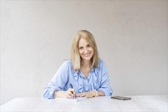 Beautiful mid-adult smiling woman is writing in her notebook while looking at camera