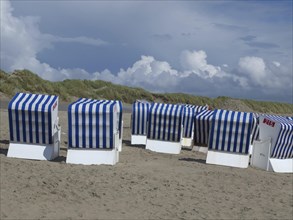 A group of blue and white beach chairs stands on the beach in front of grassy dunes and a cloudy
